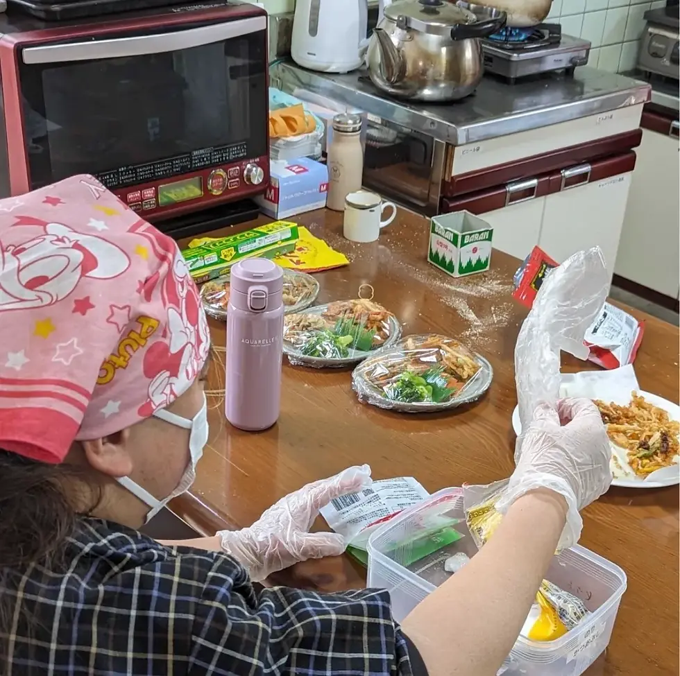 ピンクのバンダナとマスクを着けた人が、手袋をしてテイクアウトのパックに料理を詰めている様子