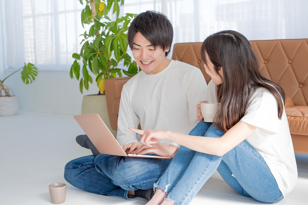 自宅のリビングで床に座り、ノートパソコンを一緒に見ながらリラックスした時間を過ごしている男女。女性はマグカップを持っており、笑顔で男性と会話しています。