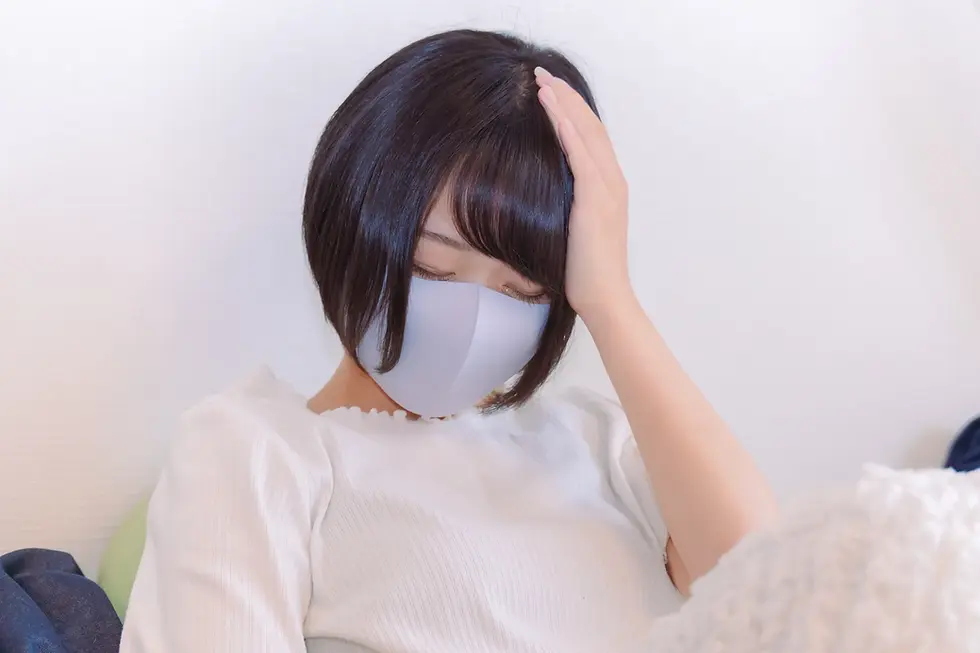 白い服を着た女性が、マスクを着用して少しうつむき、手で頭を押さえている様子