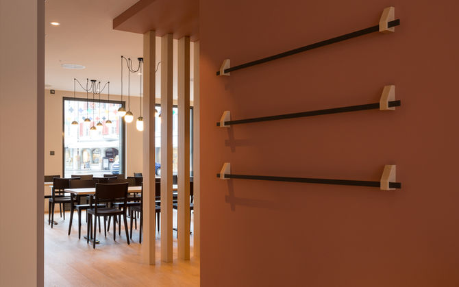 Maßgefertiger Zeitungssänder vor roséfarbener Wand, im Hintergrund Gastraum mit Pendelleuchten