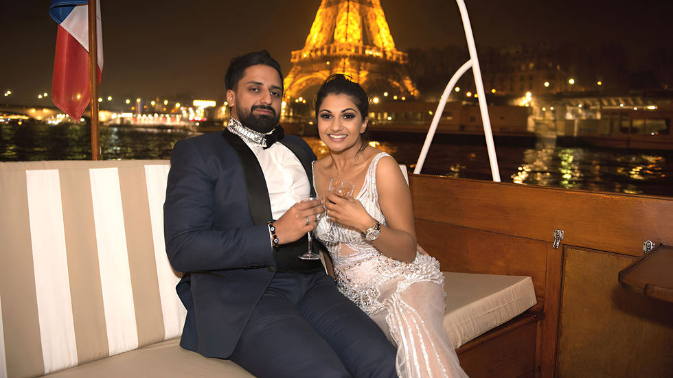 Man in a suit and woman in a white dress smiling and toasting with champagne on a boat in front of the Eiffel Tower