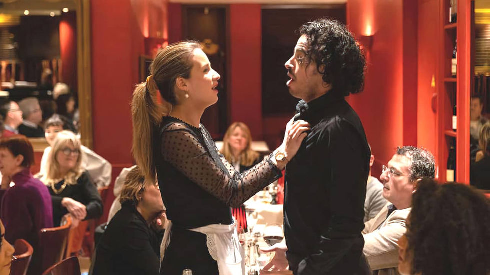 Une femme, en tenue de serveuse, et un homme se regardent et chantent dans un restaurant