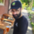 A photograph of Elliott holding a beer on the TPK patio, wearing a black dad hat with the full-color TPK backpack logo in the center of it.