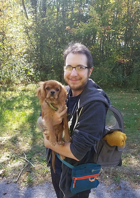 A pale skinned person with short hair and a beard holds a small ruby Cavalier King Charles Spaniel puppy in his arms while wearing a dog carrying backpack and a treatpouch.