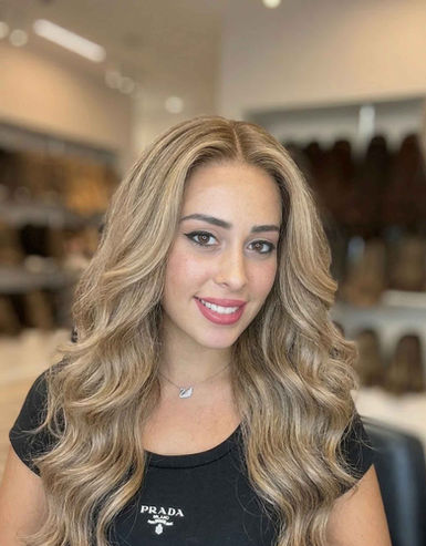 אישה צעירה מחייכת חובשת פאה ארוכה וגלית בגווני בלונד, בעלת נפח עשיר ומראה שיער טבעי.