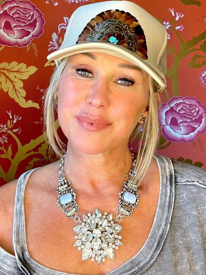 A blond woman wearing a grey sweatshirt and big statement necklace with Briley King hat