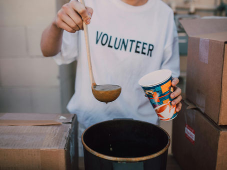 A volunteer in a white T-shirt labeled "VOLUNTEER" ladles soup into a colorful cup at a food distribution site, surrounded by cardboard boxes, highlighting community service and food assistance efforts.