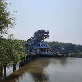 An outdoor image featuring a large dragon sculpture with a large head extending over a body of water, supported by a pier-like structure. The dragon has intricate details and appears to be made of metal, with trees flanking either side under a hazy sky.