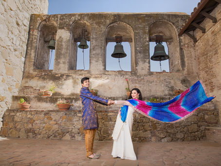 Couple in South Asian attire with a bright dupatta flying in front of a historic stone bell wall