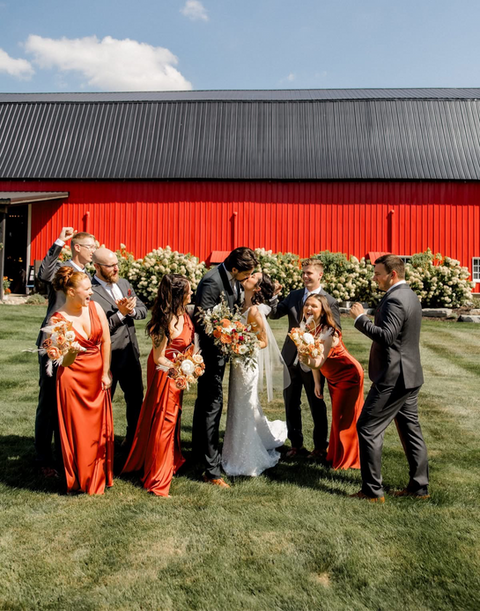 A bride and groom share a kiss in front of the barn and their wedding party