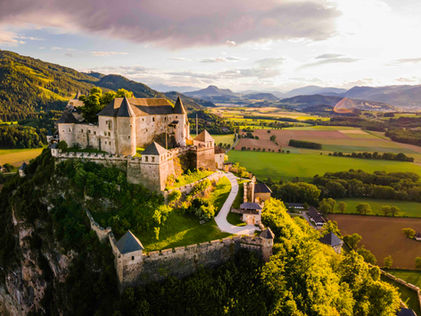 Luftaufnahme der Burg Hochosterwitz auf einem steilen Felskegel im warmen Abendlicht umgeben von grüner Landschaft.