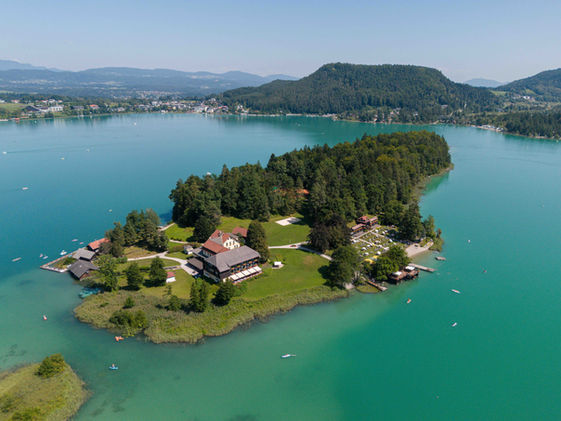 Luftaufnahme der Insel im türkisblauen Faaker See mit Hotelgebäude und Badegästen.