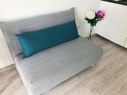 Graues Schlafsofa mit türkisfarbenem Kissen im hellen Wohnbereich der Ferienwohnung Grias Di