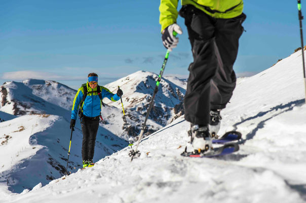 Zwei Skitourengeher steigen bei blauem Himmel über einen verschneiten Bergrücken auf.