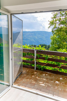 Offene Balkontür mit Blick auf den begrünten Balkon und bewaldete Berghänge im Hintergrund.