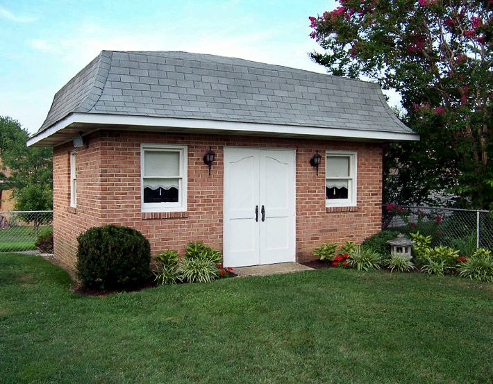 Brick shed with gray roof in a garden, flanked by shrubs and flowers. Two windows with black curtains, white double doors. Peaceful scene.