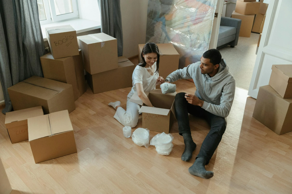 Couple unpacking boxes in a bright room with wooden floors. Labeled boxes surround them, including "Books" and "Kitchen." Smiling mood.