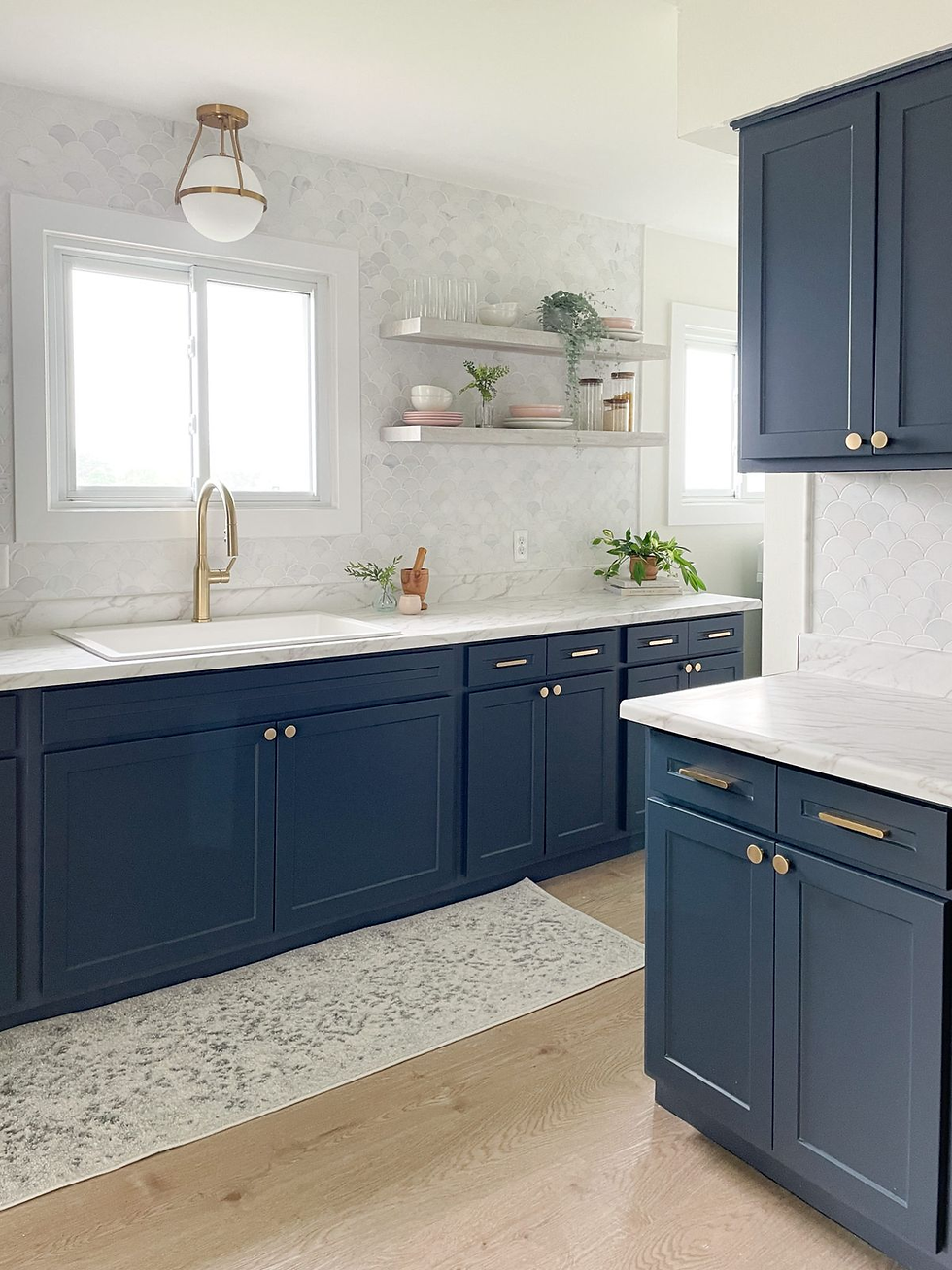 Modern kitchen with navy cabinets, gold handles, and marble countertops. Light walls, white window, potted plants, and open shelves.