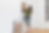 Woman in green shirt and jeans stands on ladder, hanging framed art on a white wall. Other frames lean against the wall. Bright room.
