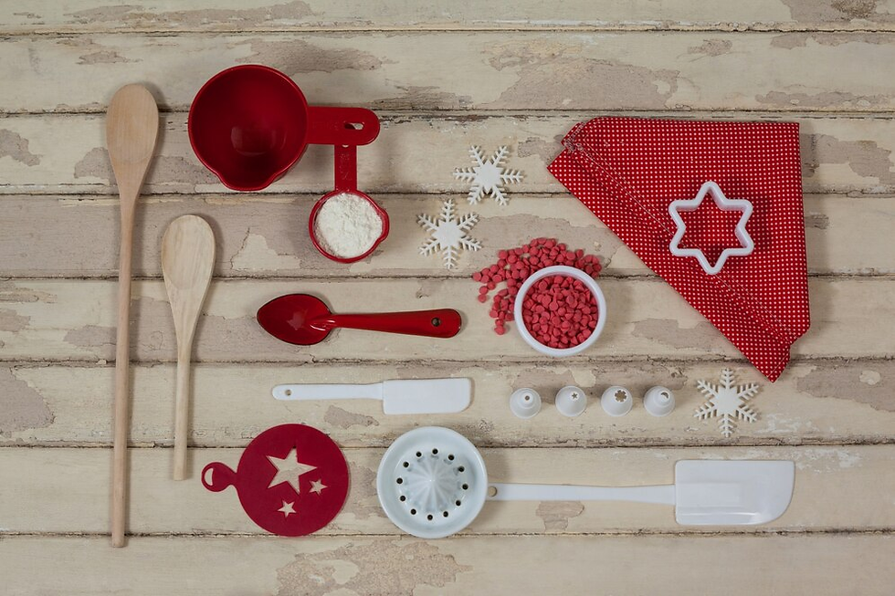 Wooden spoons, red measuring cups, a star cutter, and snowflake designs on a rustic wooden table. Festive baking items arranged neatly.