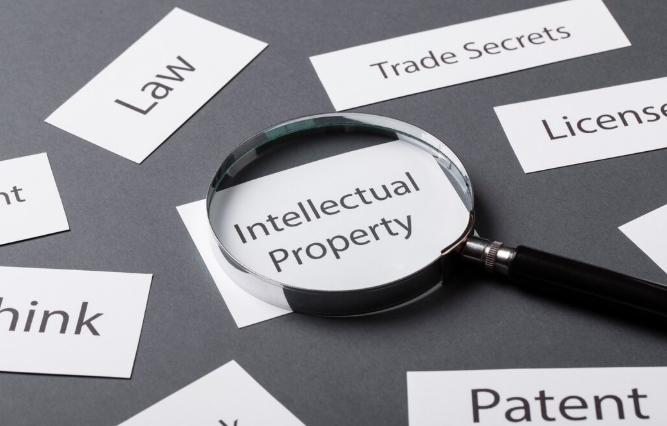Magnifying glass over "Intellectual Property" on a gray table surrounded by words "Law," "Patent," "Trade Secrets," creating a focused mood.