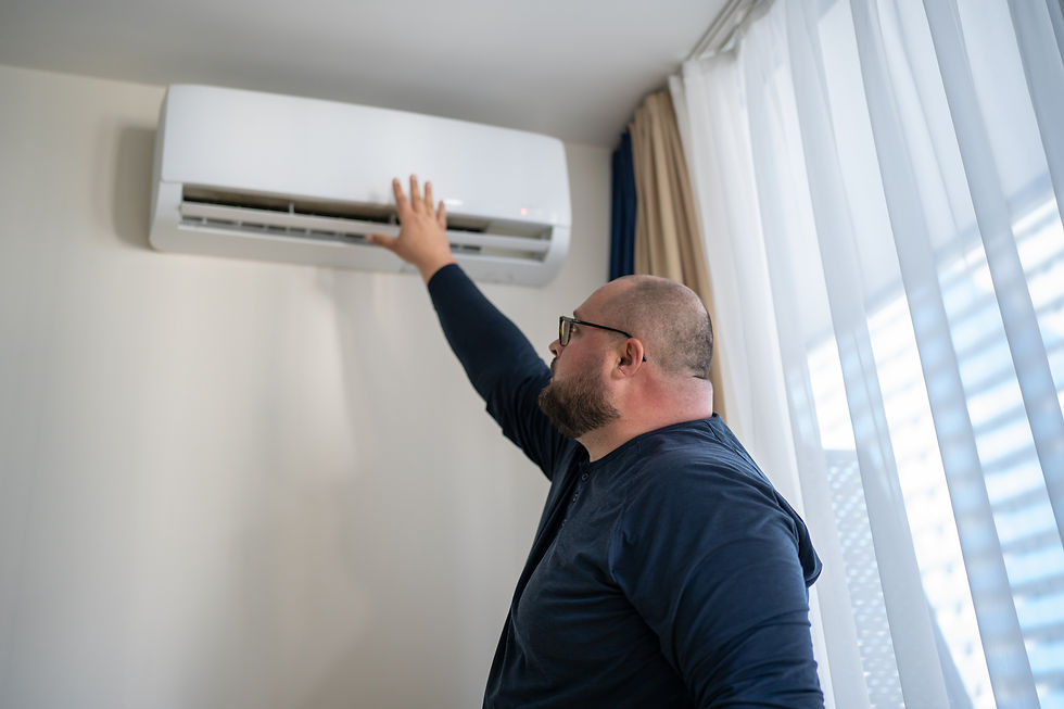 Man in a blue shirt reaches to adjust a mounted air conditioner in a bright room with sheer curtains on the right.