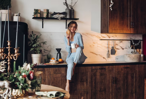 Woman in blue pajamas sits on a kitchen counter, holding a croissant and mug. Festive decor and warm lighting create a cozy atmosphere.