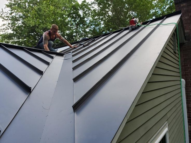 Man working on a metal roof in a green wooded area. He's adjusting equipment, wearing casual attire. Roof is sleek with visible panels.