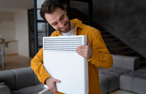 Man in a yellow jacket smiles joyfully while holding a white heater. He stands in a modern living room with gray sofas and a staircase.