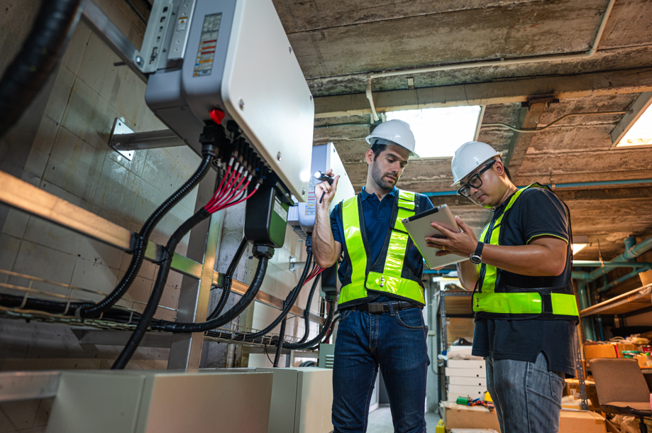 Two engineers in hard hats and reflective vests inspect electrical equipment in an industrial basement. One holds a flashlight, the other a tablet.