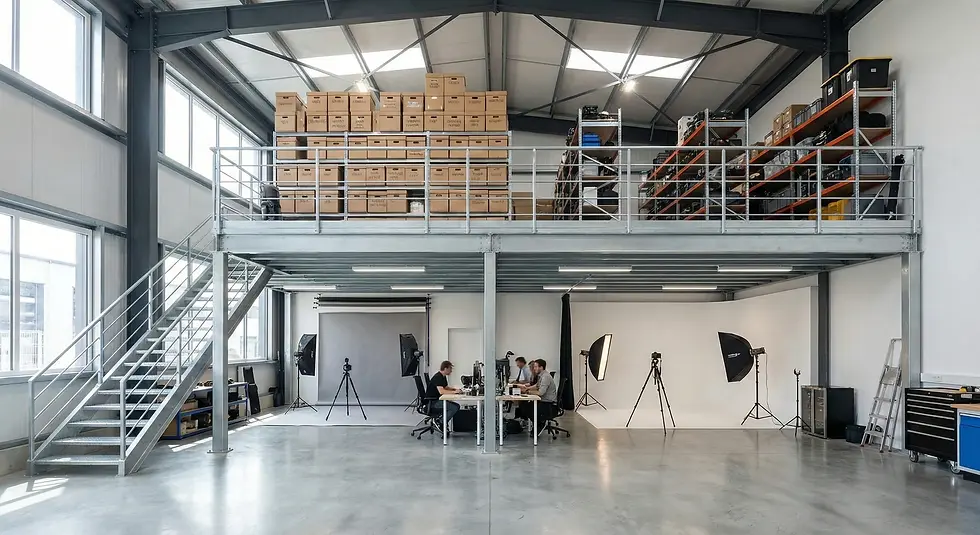 Industrial studio with mezzanine stacks of boxes, people working at a table below, and photography equipment set up. Bright, spacious atmosphere.