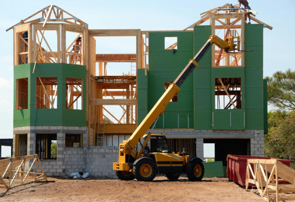 Construction site with a partially built house, green panels, and exposed wood beams. A yellow crane lifts materials against a clear sky.