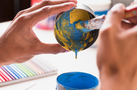 Hands painting a small bowl with blue and yellow swirls. Brushes, a blue paint pot, and colored pencils are visible in the background.