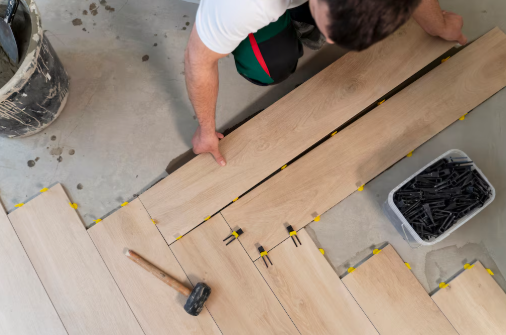 Person installing light wood laminate flooring with a mallet nearby. Floor spacers and a container of clips are visible on a concrete surface.