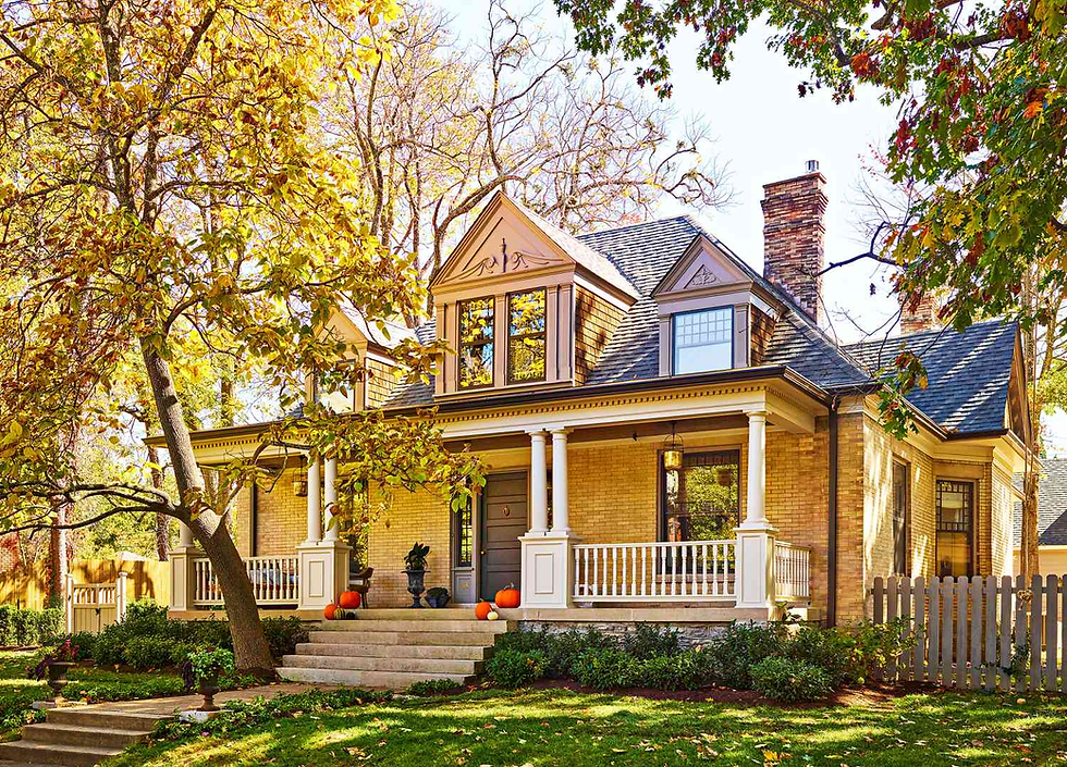 A charming two-story house with a porch, pumpkins on steps, and golden autumn leaves. Bright sunlight filters through surrounding trees.