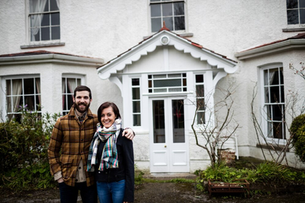 A smiling couple stands outdoors, arm in arm, in front of a white house with large windows. He wears a plaid jacket; she's in a patterned scarf.