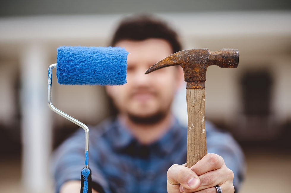 Man holding a blue paint roller and a rusty hammer in focus, wearing a plaid shirt. Background is blurred, creating a neutral mood.