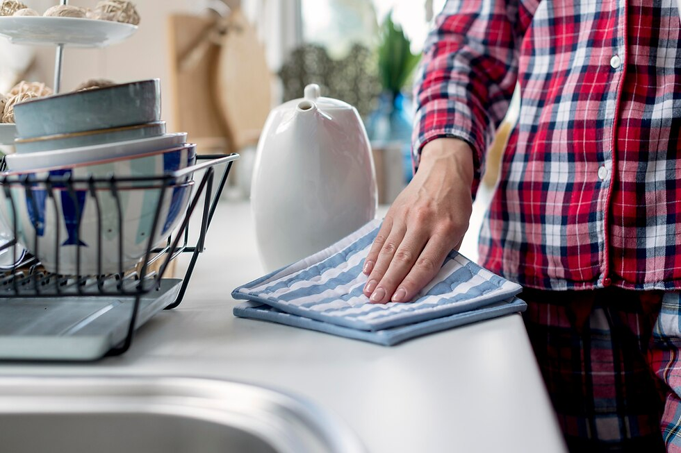 Person in red plaid pajamas wipes a countertop with a striped cloth. A dish rack with bowls and a white teapot are nearby. Bright kitchen.