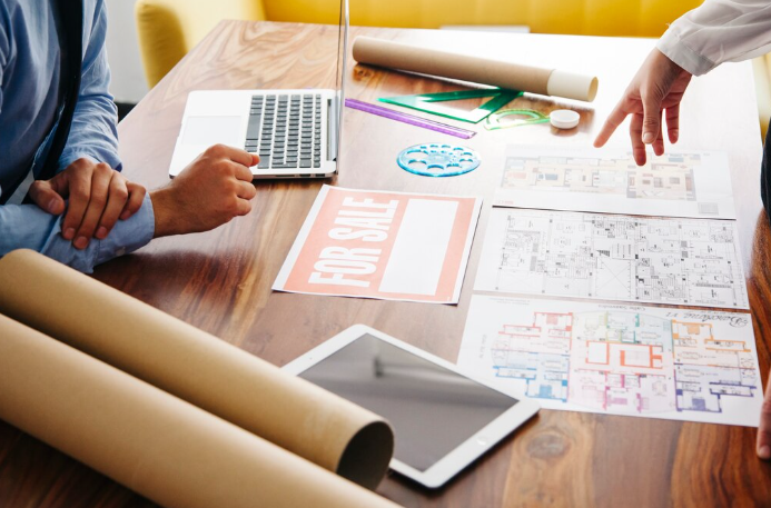 Two people at a wooden table with blueprints, a laptop, and a "For Sale" sign. One points at plans; an iPad and rolled papers are visible.