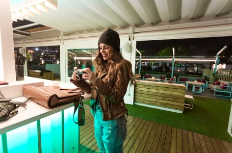 Woman in a brown jacket and beanie smiles while holding a camera inside a lit-up, modern outdoor lounge with wooden deck and green seating.