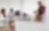 Man in apron chops vegetables in modern kitchen; couple sits at counter with drinks, discussing. Bright, minimalist setting.