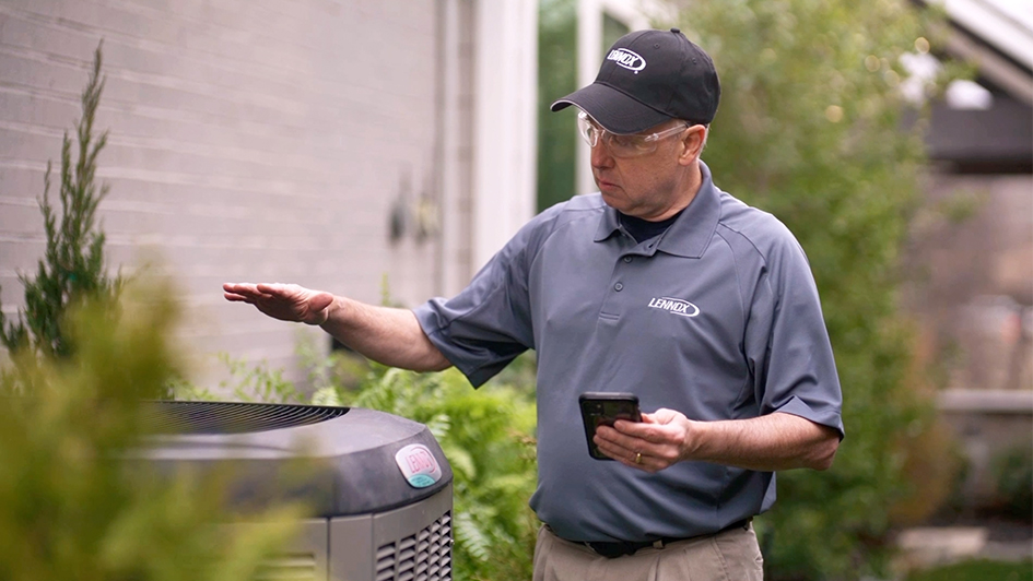 Man in Lennox uniform checks an outdoor HVAC unit, holding a phone. He's focused, standing near a brick wall with greenery around.