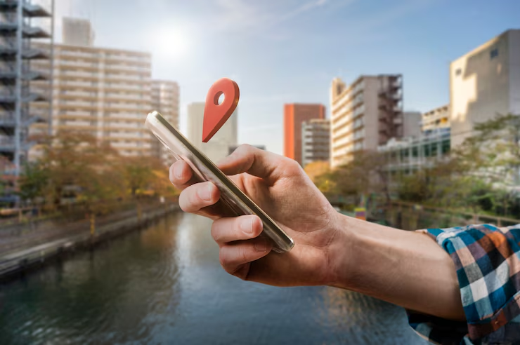 Hand holding a smartphone with a floating map pin icon. City buildings and river in the background. Bright, sunny day; plaid shirt visible.