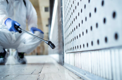 Person in protective suit and blue gloves spraying a surface in an industrial setting. Focus on the nozzle, metal wall, and tiled floor.