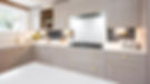 Bright kitchen with gray cabinets, gold handles, and white countertops. Gas stove, toaster, and flowers next to window. Cozy and tidy.