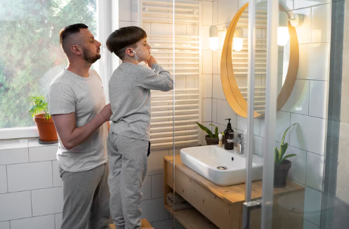 Father and son in pajamas brushing teeth in a modern bathroom with plants and wooden elements. Sunlight streams through the window.