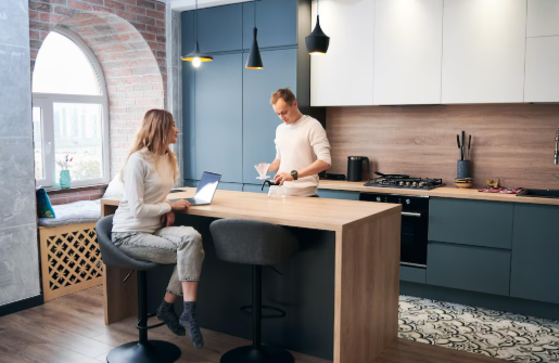 Man and woman in modern kitchen, she sits on stool with laptop, he stands with mug. Bright, cozy room with blue and white cabinets.