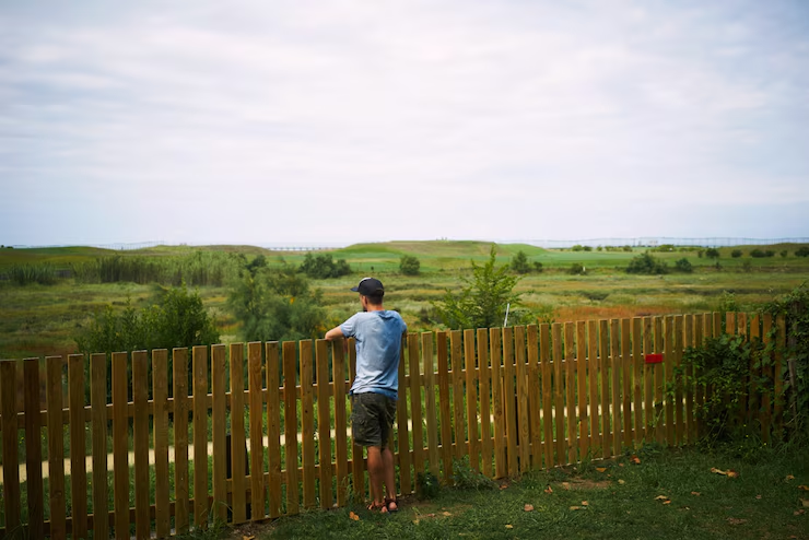 Man in blue shirt and cap leans on wooden fence, gazing at lush green landscape under cloudy sky, evoking a serene mood.