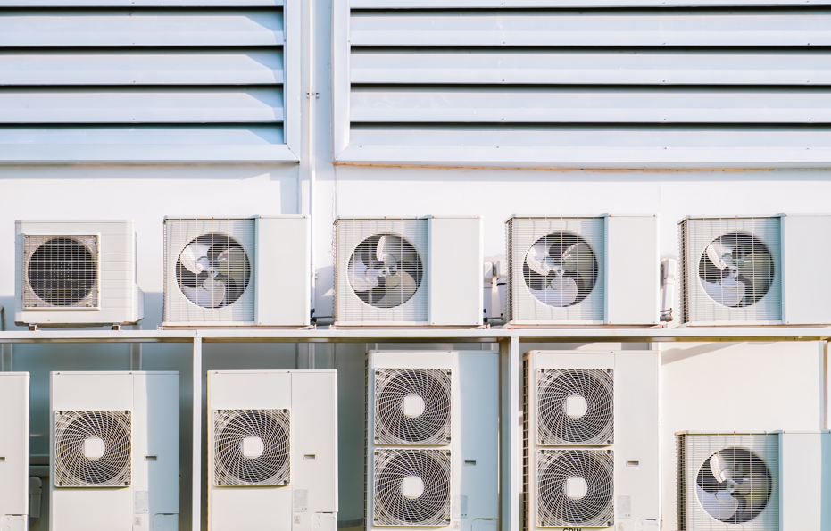 Outdoor view of multiple white air conditioning units mounted on a building wall with horizontal vents. Bright lighting and no visible text.