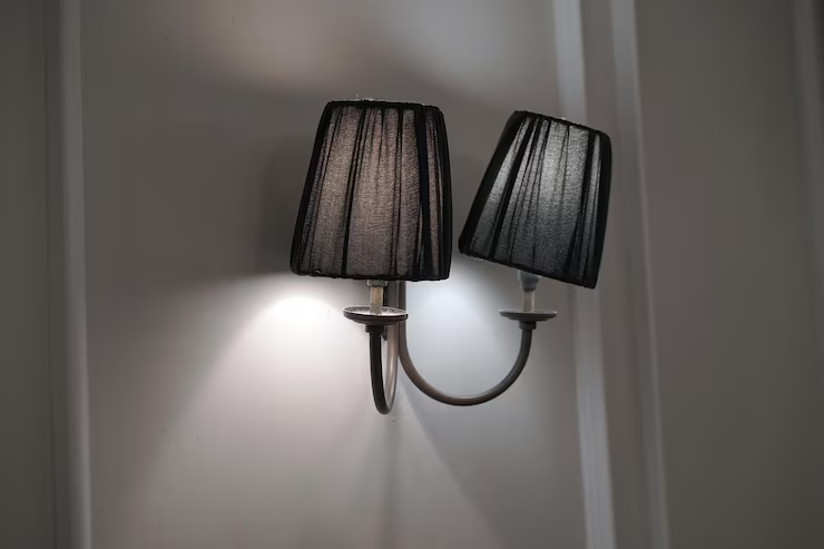 Wall-mounted light fixture with two black fabric lampshades emits soft light. Set against a gray wall, creating an elegant, understated ambiance.